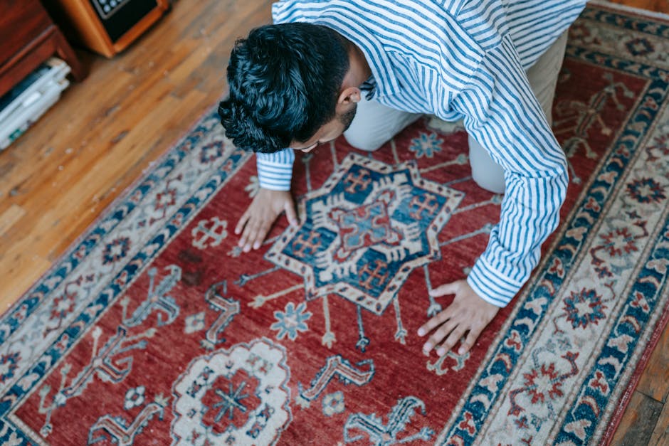 A person kneeling on a red patterned rug with intricate blue, white, and beige designs, using their hands to clean or inspect the surface. The rug is situated on a hardwood floor near a wooden bookshe