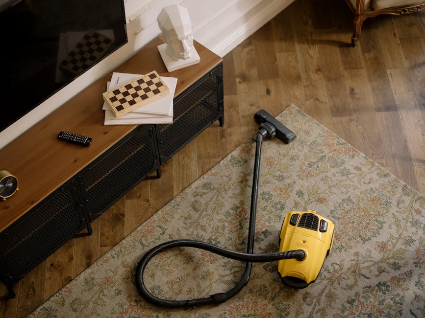 A yellow handheld vacuum cleaner with a flexible hose attachment resting on a light-colored patterned rug in a living room. The room features a wooden floor and a wooden sideboard with black metal fra