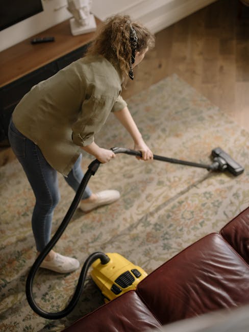 A yellow handheld vacuum cleaner with a flexible hose attachment resting on a light-colored patterned rug in a living room. The room features a wooden floor and a wooden sideboard with black metal fra